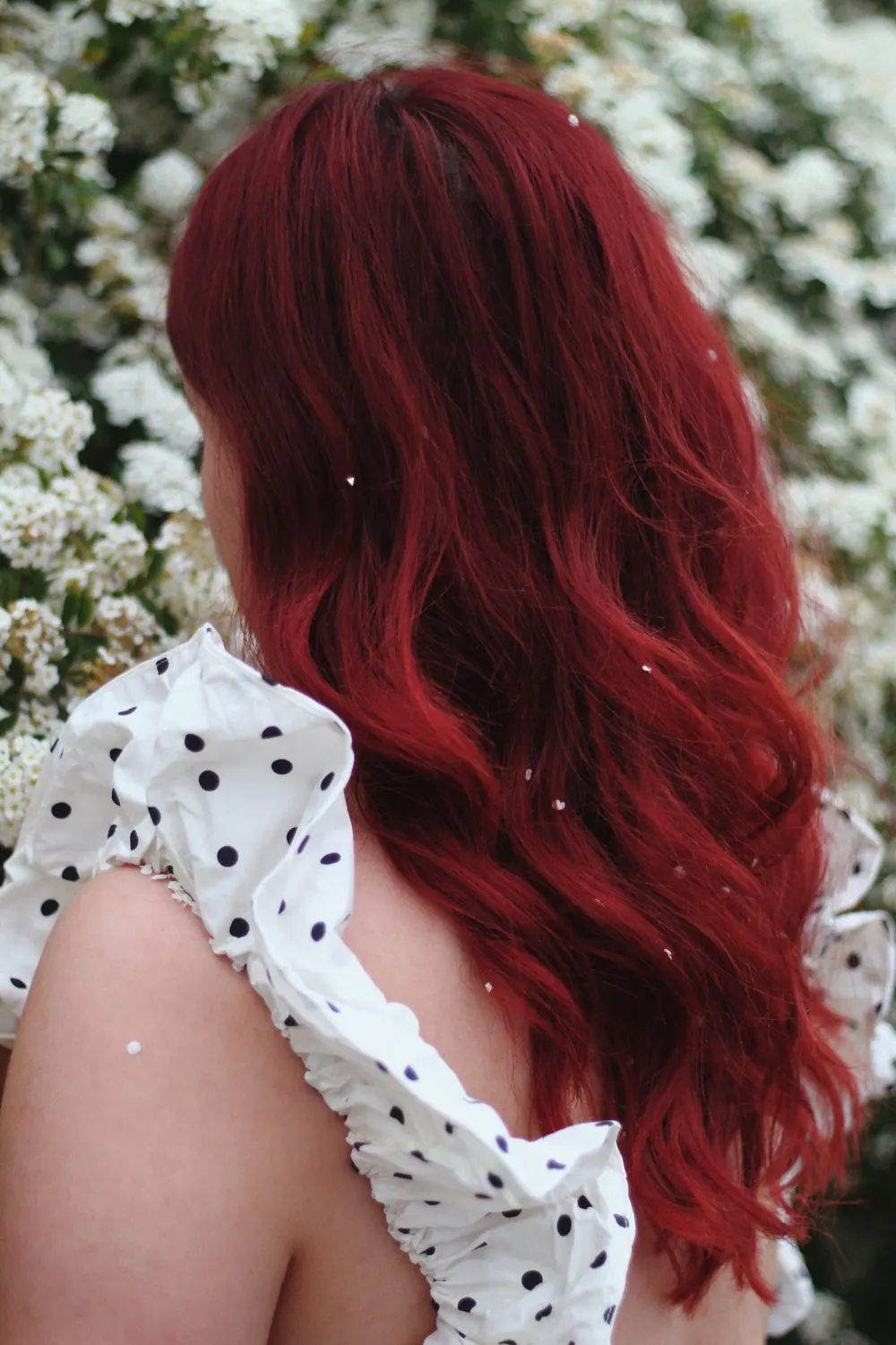 Back view of a woman with long red hair in a polka dot dress standing near white flowers.