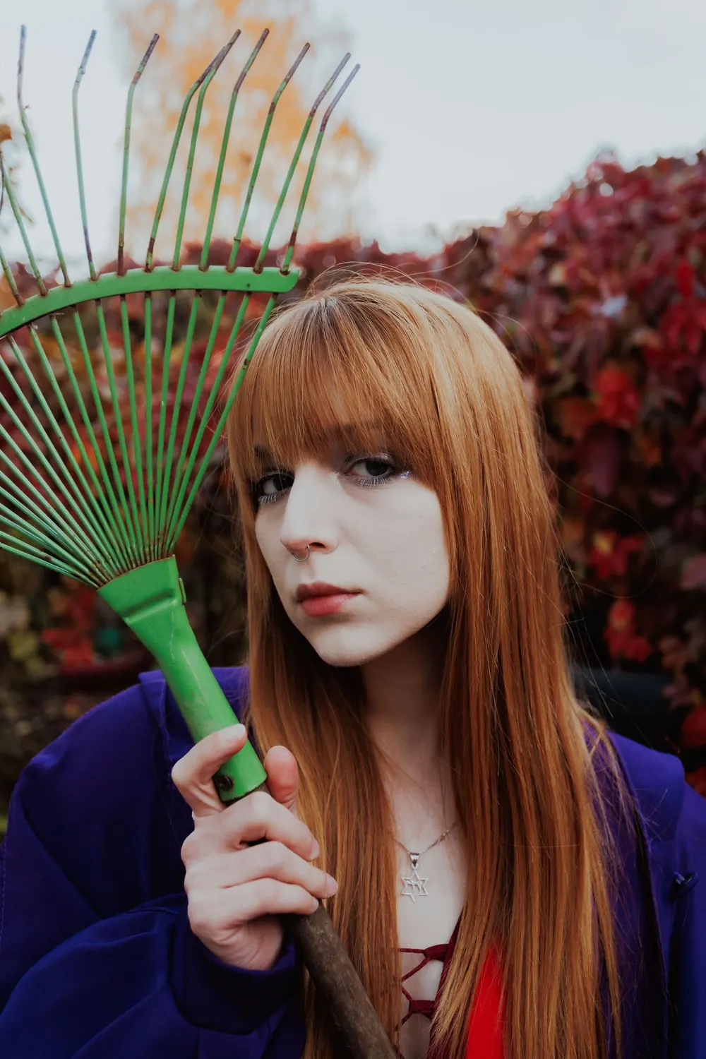 Red-haired woman with green rake, surrounded by autumn foliage, showcasing gardening theme.
