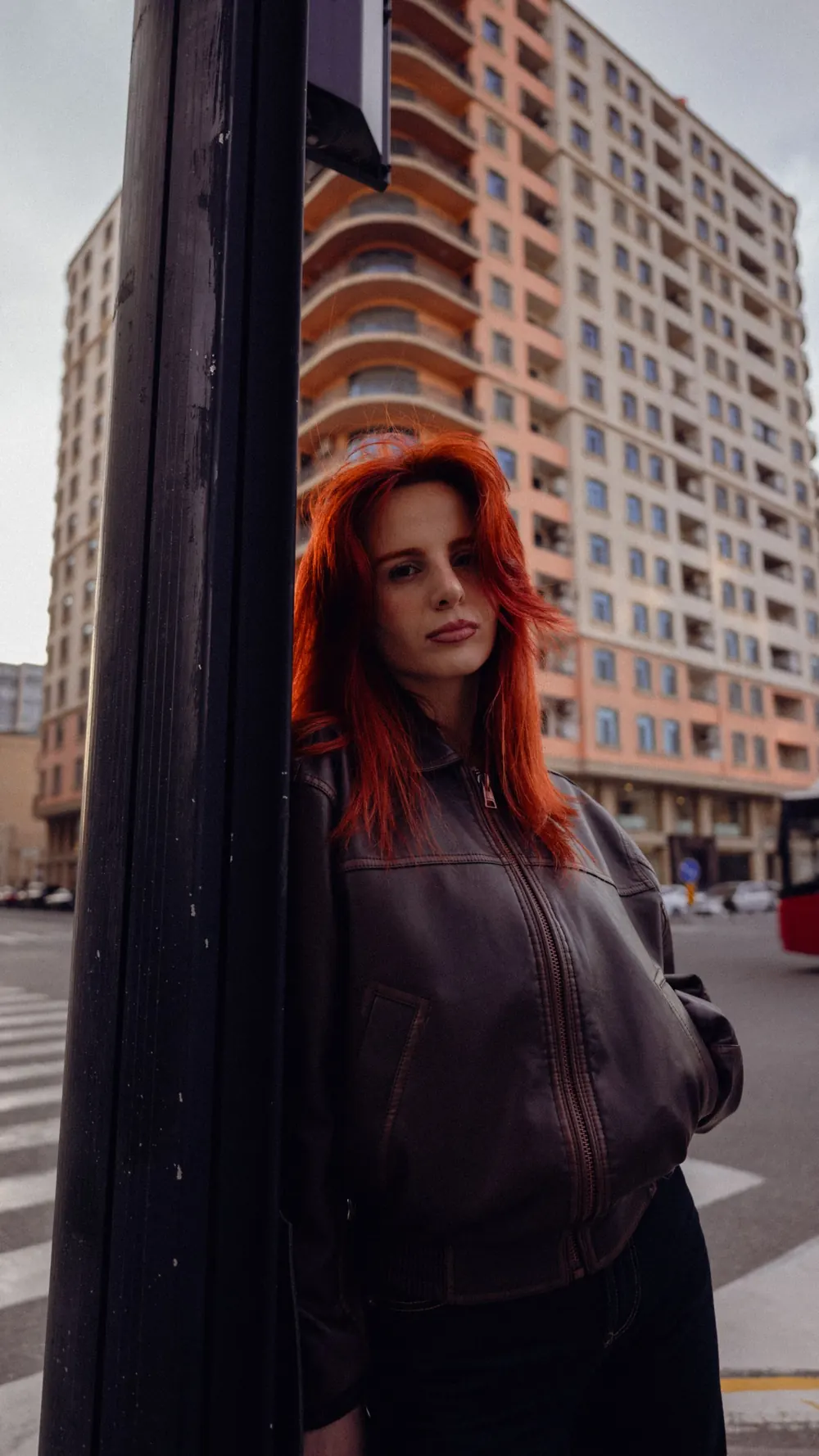Woman with red hair poses by a city street, building in background.
