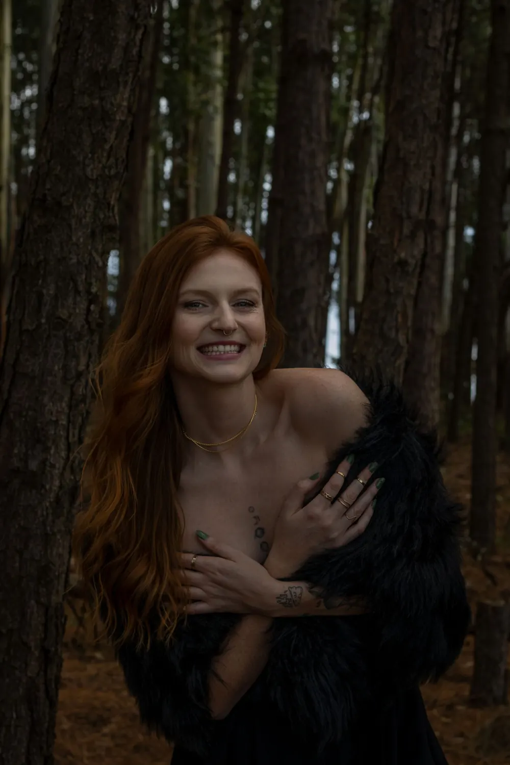 Red-haired woman in black fur wrap smiling amidst forest trees.