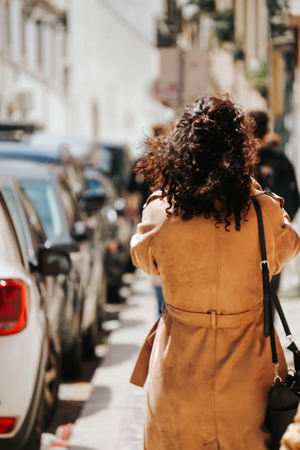 Brunette woman in a coat walking along a city sidewalk with cars parked beside.