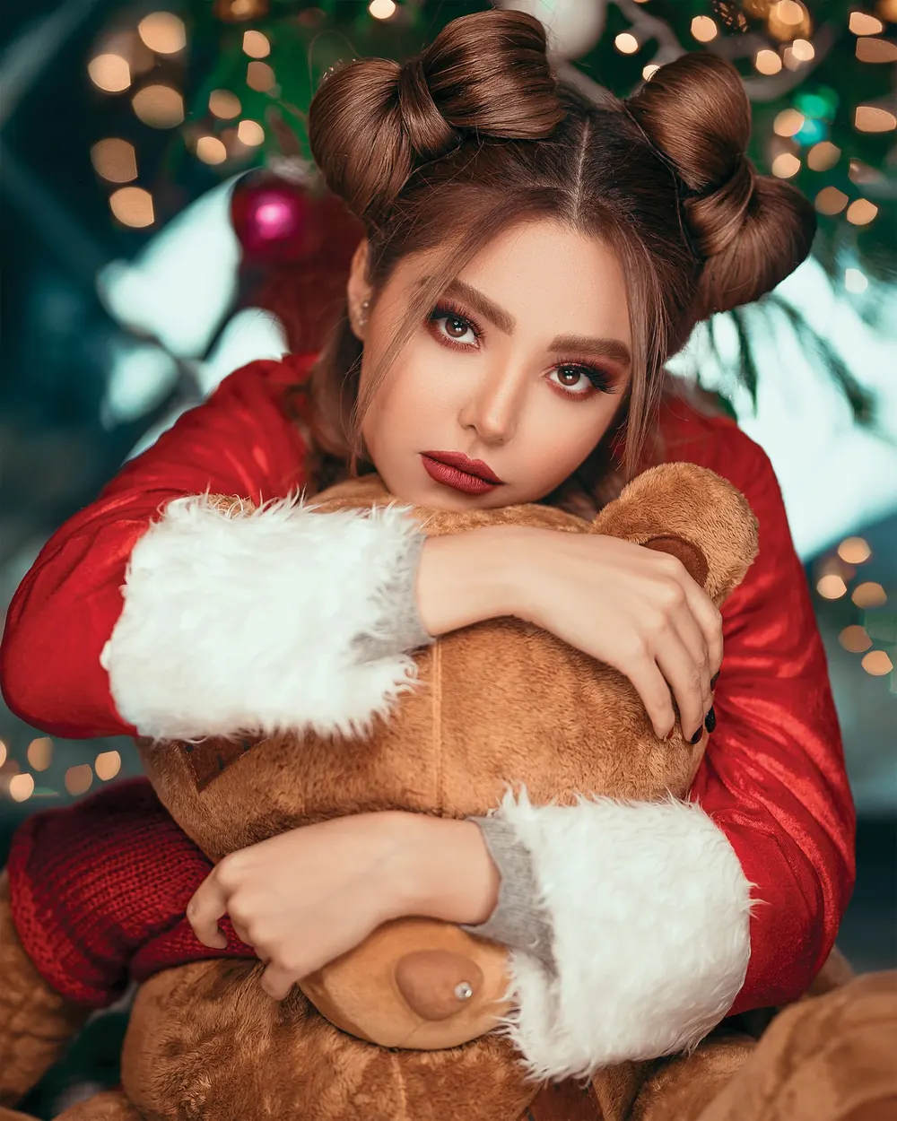 Beautiful woman in red Santa costume hugging a teddy bear with blurred Christmas lights.