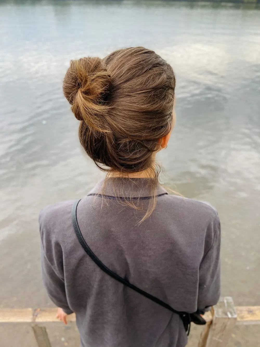 Back view of a woman with a bun hairstyle, wearing a jacket, looking at a calm body of water outdoors.