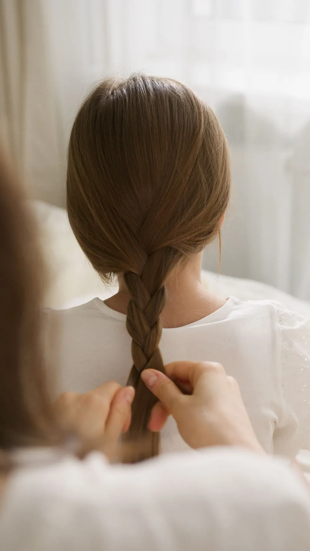 Child's hair being braided by adult hands indoors, creating a delicate hairstyle.
