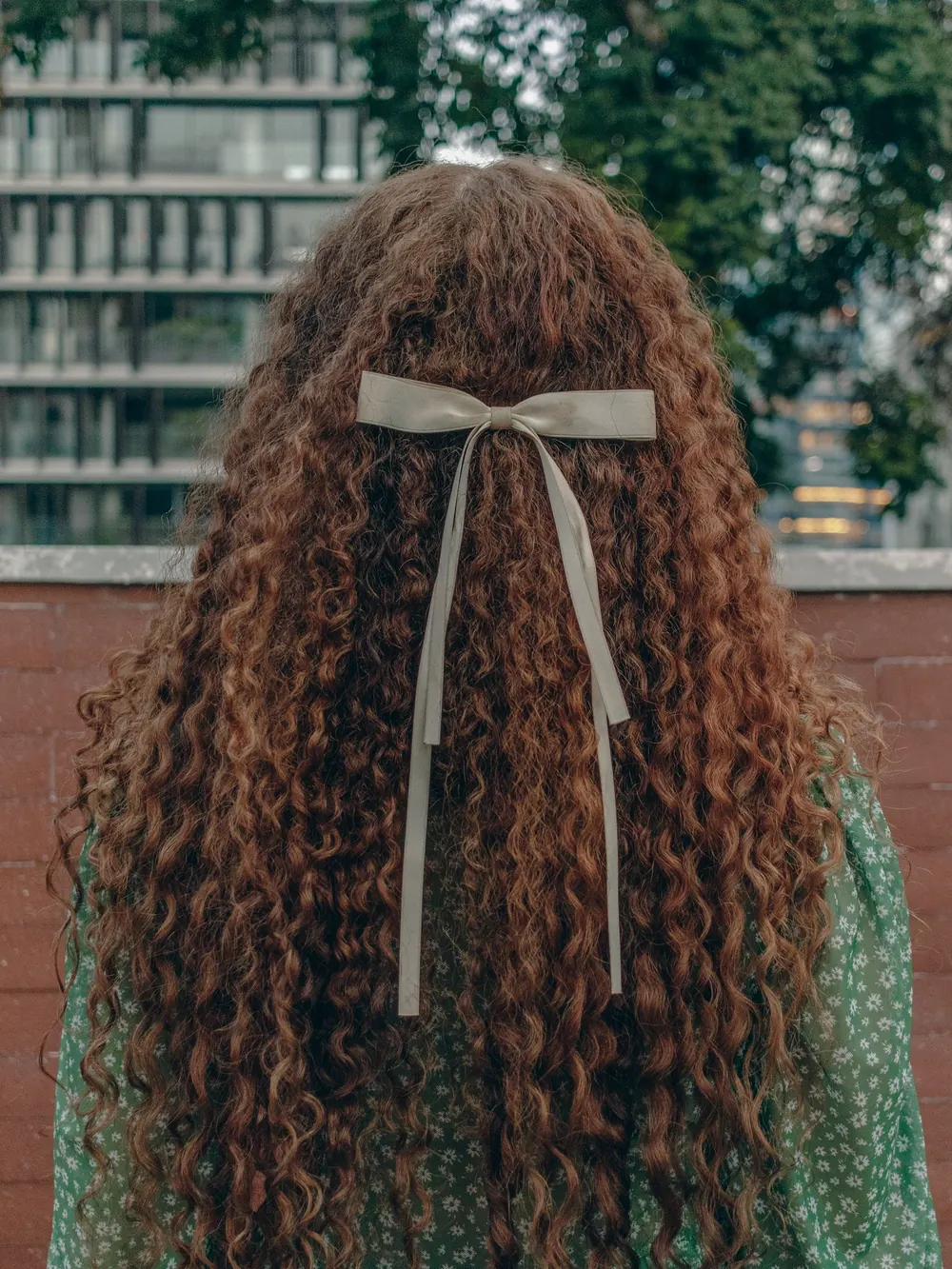 A woman with long curly hair tied with a bow, standing outdoors in a city setting.