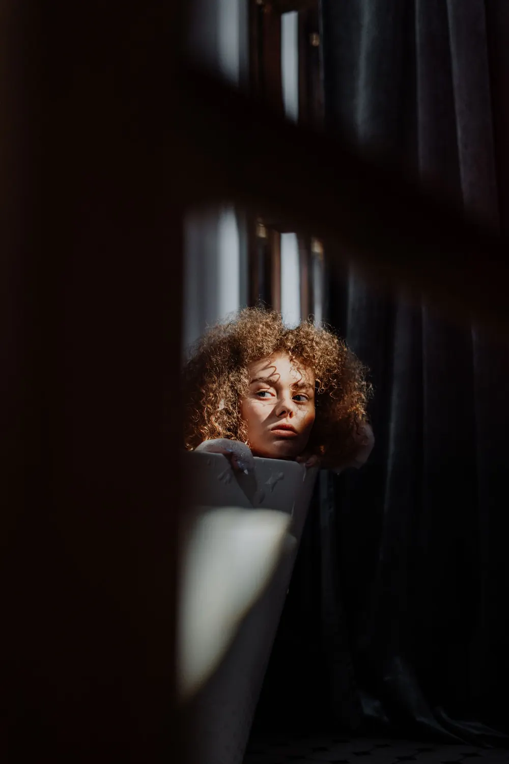 Young woman with curly hair relaxing in a sunlit bathtub indoors.