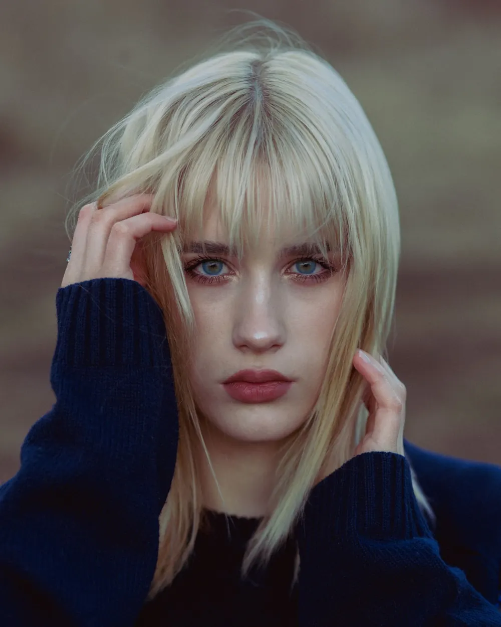 Close-up portrait of a young blonde woman with fringe and blue eyes, captured outdoors.