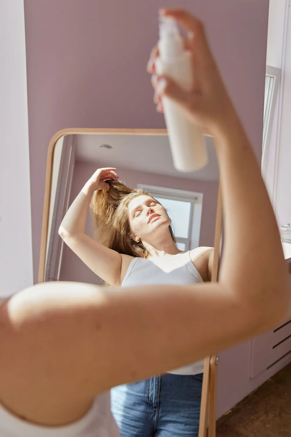 Woman using hair spray in front of a mirror, enjoying a beauty routine in natural daylight.